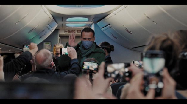A masked Alexei Navalny waves as he boards a plane while passengers take photos and videos of him.