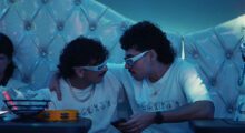 Two men, dressed identically and wearing white paper glasses, look at one another.